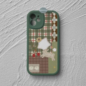 May include: A green phone case with a patchwork design featuring a plaid pattern, a white star, a bunny, cherries, and a flower. The case has a green border and a camera lens protector.