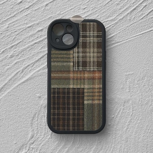 May include: A black smartphone case featuring a patchwork design of various plaid fabric patterns in shades of brown, tan, olive green, and cream. The case has a black camera cutout frame and a small silver metallic tab at the top center edge.