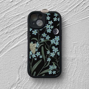 以下が含まれることがあります： 黒い携帯電話ケース。青いワスレナグサの花と蛾が暗い背景に描かれた花柄のデザインです。