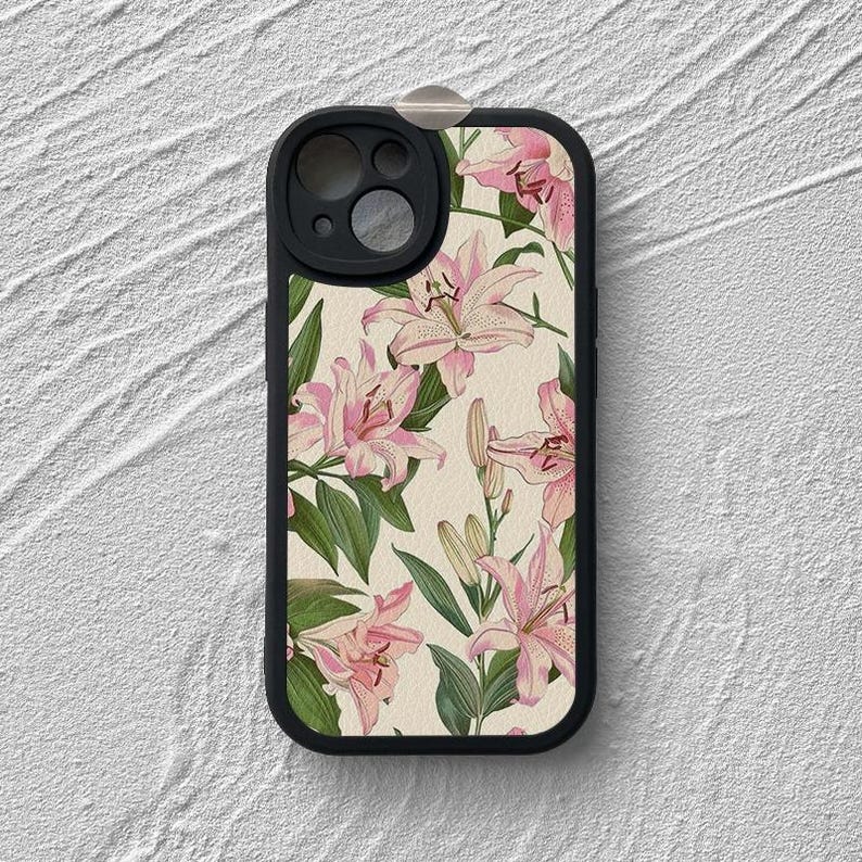 May include: A black phone case with a floral design featuring pink lilies on a cream background. The case has a cutout for the camera lens.