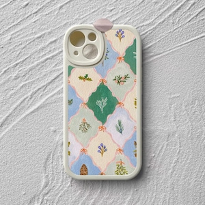 Op de afbeelding: Een telefoonhoesje met een crèmekleurige rand en een decoratief patroon. Het patroon heeft een herhalend ontwerp van ruitvormen in groen, blauw en crème, met bloemen en botanische illustraties en roze strikken. Bovenop is een kleine, roze lus bevestigd.
