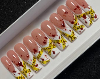 May include: A set of ten long, coffin-shaped press-on nails featuring a French manicure design with a nude base and white tips. The tips are decorated with yellow caution tape patterns reading CAUTION and blood-like red splatter accents.
