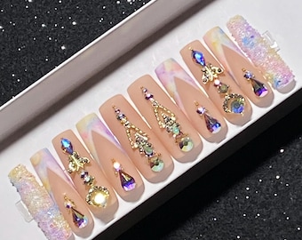 May include: A set of long, square-shaped false nails. Each nail is a nude color with a white French tip and adorned with rhinestones and gold-colored embellishments. Some nails feature dangling gems and iridescent glitter.
