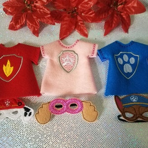 May include: Three felt costumes for a doll or action figure, featuring a red shirt with a yellow and orange flame, a pink shirt with a silver paw print, and a blue shirt with a gray paw print.  Three felt masks are also included, one with a red and white paw print, one with pink sequins, and one with a brown and black paw print.