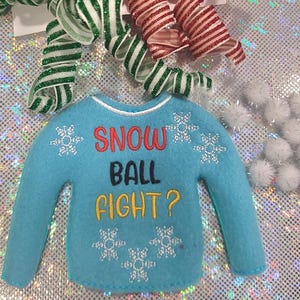 May include: A light blue felt sweater with the text "SNOW BALL FIGHT?" in red, black, and yellow. White snowflake designs are embroidered on the sweater. Green and red striped ribbons and white pom-poms are also visible.