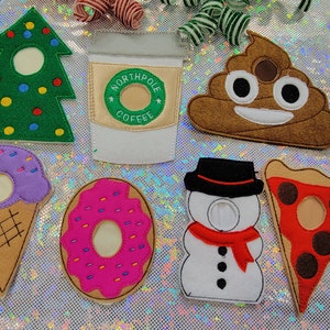 May include: A collection of felt food and holiday-themed items, including a green Christmas tree, a coffee cup with "Northpole Coffee" text, a smiling poop emoji, an ice cream cone, a pink donut, a snowman, and a pizza slice.