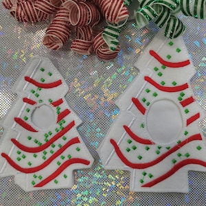May include: Two white Christmas tree-shaped ornaments with red and green accents. The ornaments have a hole in the center and are decorated with red stripes and green squares. Red and green striped ribbons are in the background.
