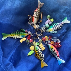 May include: A collection of colorful fish-shaped keychains. Each keychain features a unique design with vibrant colors, including red, yellow, green, and blue. They are adorned with decorative beads and charms, arranged on a blue fabric background.