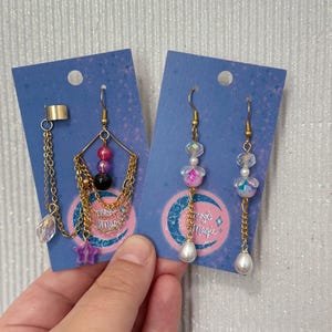 May include: Two pairs of handmade earrings on blue cards. One pair features a gold ear cuff with a chain and a clear crystal. The other pair has pink and clear beads with a dangling pearl. The cards have a moon and stars design.