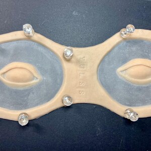 May include: A beige-colored eye mask with two eye-shaped openings, each featuring a light-colored, lip-shaped design. Clear, rounded studs are placed around the perimeter. The text "SSTUS" is visible on the mask.