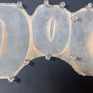 May include: A light-colored, irregularly shaped object with three connected oval sections, each featuring a central, elongated slit. Clear, rounded pins are placed around the perimeter. The object rests on a dark surface.