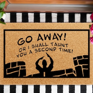 May include: A rectangular doormat with a light brown surface and a black border. The mat features the text "GO AWAY! OR I SHALL TAUNT YOU A SECOND TIME!" in black, along with a silhouette of a person and brick walls.