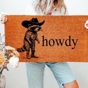 May include: A rectangular doormat with a raccoon wearing a cowboy hat and boots, with the word "howdy" in black. The mat is a natural brown color, and the raccoon illustration is in black.