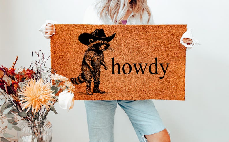 May include: A rectangular doormat with a raccoon wearing a cowboy hat and boots, with the word "howdy" in black. The mat is a natural brown color, and the raccoon illustration is in black.