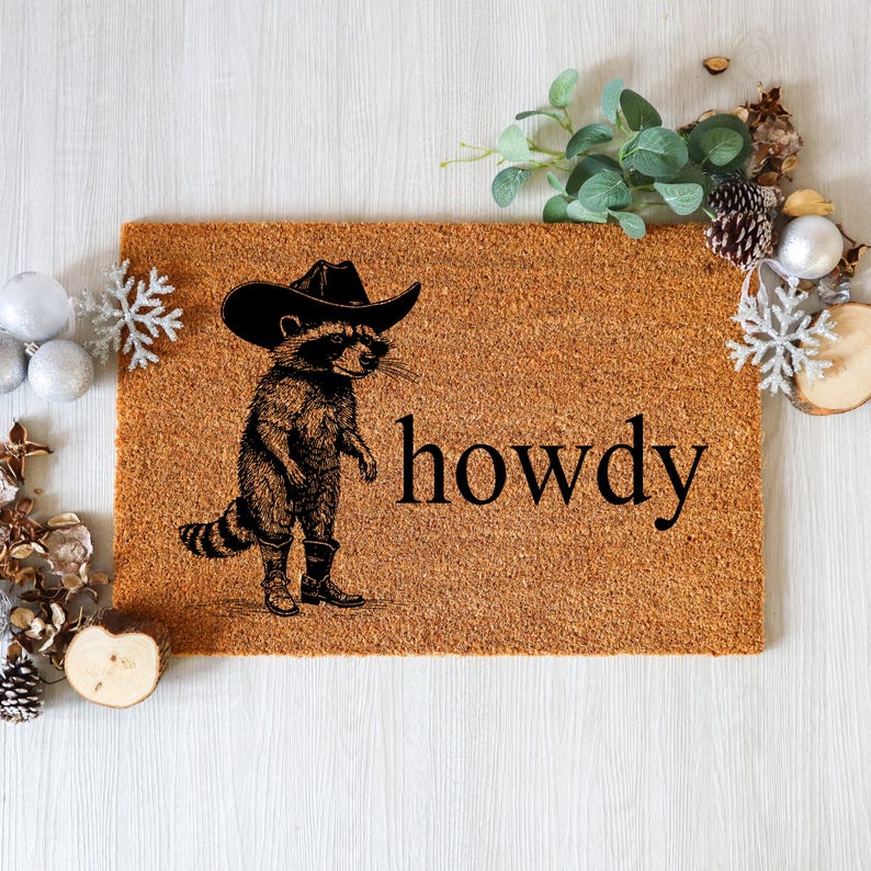 May include: A rectangular doormat with a brown coir surface. A black and white illustration of a raccoon wearing a cowboy hat and boots is on the left. The word "howdy" is printed in black on the right.