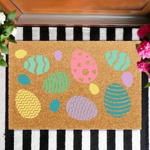 May include: A rectangular doormat made of natural fibers, featuring colorful Easter egg designs in shades of pink, teal, yellow, and purple. The mat is placed on a black and white striped surface, creating a festive and welcoming entrance.