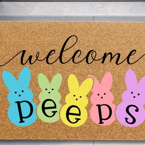 May include: A rectangular doormat with a light brown background. The word "welcome" is written in black cursive script. Below are five colorful bunny shapes in blue, green, yellow, pink, and purple, with the word "peeps" in black.