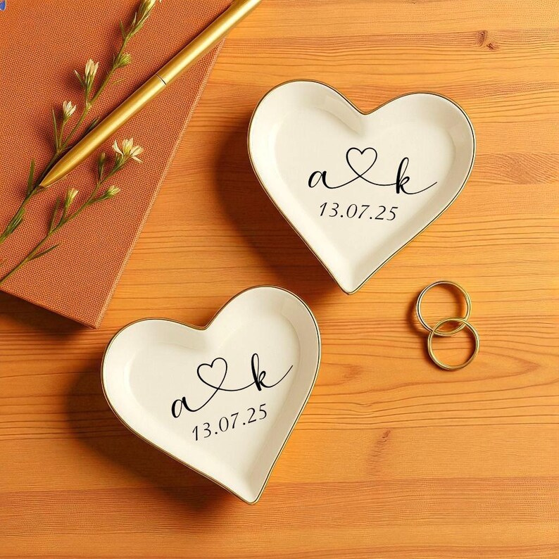 May include: Two heart-shaped white ceramic ring dishes with gold trim, each inscribed with initials and a date in black script. Gold wedding rings and a gold pen rest on a wooden surface, alongside a small notebook and sprigs of flowers.