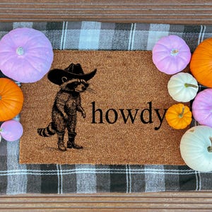May include: A brown doormat with a raccoon wearing a cowboy hat and boots, with the word "howdy" printed on it. The mat is surrounded by various pumpkins in shades of orange, white, and purple, creating a fall theme.