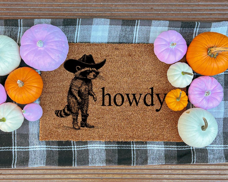 May include: A brown doormat with a raccoon wearing a cowboy hat and boots, with the word "howdy" printed on it. The mat is surrounded by various pumpkins in shades of orange, white, and purple, creating a fall theme.