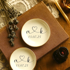 May include: Two small, round, off-white dishes with gold rims. Each dish has the initials "a & k" with a heart symbol and the date "13.07.25" in black script. The dishes rest on a brown book, with a feather and dried flowers nearby.