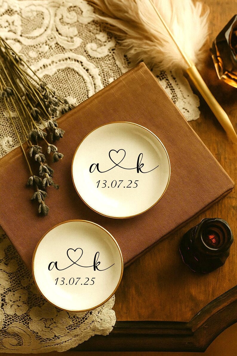 May include: Two small, round, off-white dishes with gold rims. Each dish has the initials "a & k" with a heart symbol and the date "13.07.25" in black script. The dishes rest on a brown book, with a feather and dried flowers nearby.