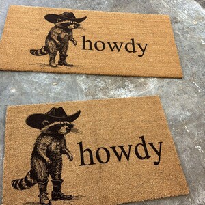 May include: Two rectangular doormats made of natural fiber. Each mat features a raccoon wearing a cowboy hat and boots, with the word "howdy" printed in black. The mats are placed on a gray surface.