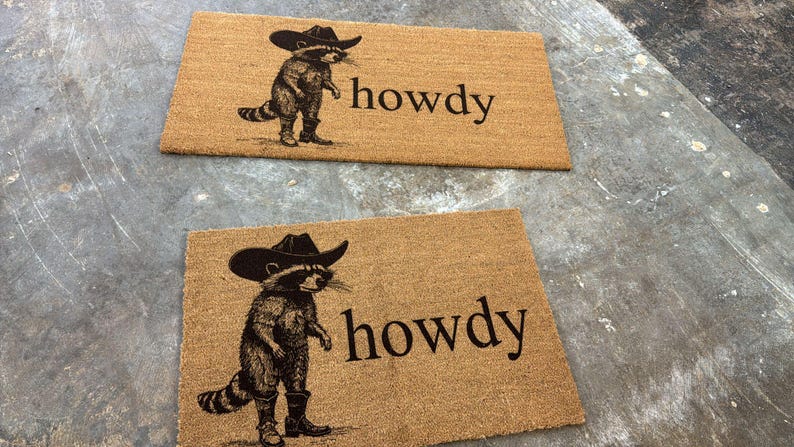 May include: Two rectangular doormats made of natural fiber. Each mat features a raccoon wearing a cowboy hat and boots, with the word "howdy" printed in black. The mats are placed on a gray surface.