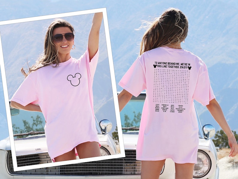 May include: A pale pink t-shirt with a black Mickey Mouse outline on the front. The back of the shirt features a word search puzzle with the text "TO ANYONE BEHIND ME, WE'RE IN THIS LINE TOGETHER. ENJOY!"
