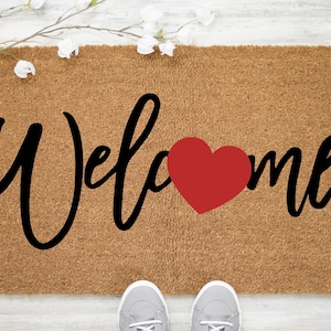 May include: A rectangular doormat made of natural fibers, featuring the word "Welcome" in black script with a red heart replacing the "o". White flowers and a pair of gray sneakers are visible.