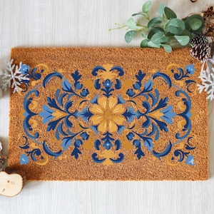 May include: A rectangular doormat with a brown coir base and a symmetrical floral design in shades of blue and yellow. The design features a central flower and ornate scrollwork. Decorative elements like snowflakes and pinecones surround the mat.