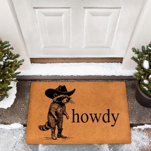May include: A rectangular doormat with a raccoon wearing a cowboy hat and boots. The raccoon is standing next to the word "howdy" in black text. The mat is brown and sits in front of a white door, with two potted evergreen trees on either side.