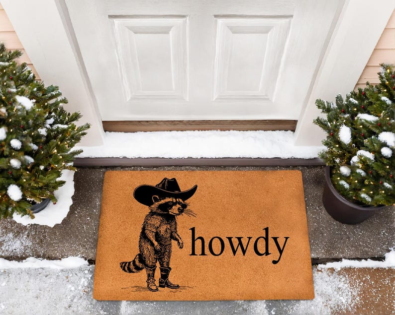 May include: A rectangular doormat with a raccoon wearing a cowboy hat and boots. The raccoon is standing next to the word "howdy" in black text. The mat is brown and sits in front of a white door, with two potted evergreen trees on either side.
