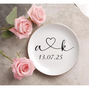 May include: A white ceramic plate with a gold rim features the intertwined letters "a & k" and a heart symbol, along with the date "13.07.25" in black script. Three pink roses with green leaves are arranged near the plate.