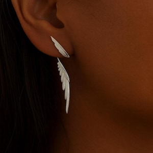 Puede incluir: Pendientes de plata con forma de pluma. Uno es una pequeña pluma simple, el otro cuelga con un diseño de pluma detallado y en capas. Los pendientes se llevan en una oreja, destacando su apariencia delicada y elegante.