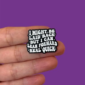 Może przedstawiać: Czarno-biały urok do butów z napisem "I MIGHT BE LAID BACK BUT I CAN LEAN FORWARD REAL QUICK" w stylu retro. Urok jest trzymany w dłoni na fioletowym tle.