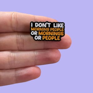 May include: A black rectangular charm with the text "I DON'T LIKE MORNING PEOPLE OR MORNINGS OR PEOPLE" in white and orange lettering. The charm is held in a hand against a light purple background.