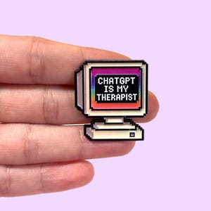 Op de afbeelding: Een pixelachtige computermonitor pin met de tekst "ChatGPT IS MY THERAPIST" op het scherm. De monitor heeft een witte rand en een regenboogkleurige achtergrond. De pin wordt tegen een lichtpaarse achtergrond gehouden.