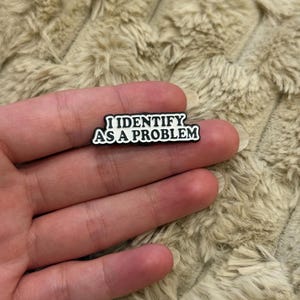 Puede incluir: Un pequeño dije rectangular en blanco y negro con el texto "I IDENTIFY AS A PROBLEM". El dije está sobre una mano, con un fondo beige suave y texturizado.