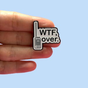 Puede incluir: Un pin de esmalte blanco y negro con forma de walkie-talkie con las palabras "WTF, over." El pin tiene forma rectangular con una pequeña antena y un diseño detallado de los botones y la pantalla del walkie-talkie.