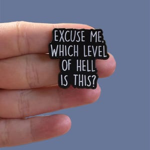 Puede incluir: Pin de esmalte negro con el texto blanco "EXCUSE ME, WHICH LEVEL OF HELL IS THIS?". El pin tiene forma rectangular con esquinas redondeadas y un borde negro. El fondo es azul liso.