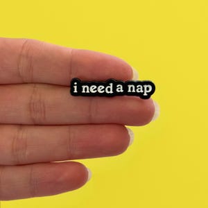 Può includere: Spilla rettangolare in bianco e nero con la scritta bianca "i need a nap". La spilla è tenuta in mano su uno sfondo giallo brillante.