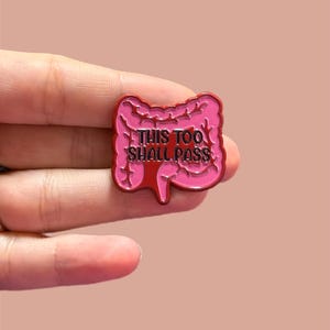 以下が含まれることがあります： 白い文字で「THIS TOO SHALL PASS」と書かれた、腸の形をしたピンクのエナメルピン。明るいピンク色の背景に、2本の指でピンが挟まれています。