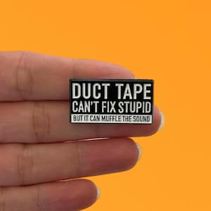 May include: A rectangular black and white enamel pin with the text "DUCT TAPE CAN'T FIX STUPID BUT IT CAN MUFFLE THE SOUND". The pin is set against an orange background.