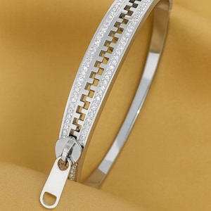 May include: A silver-toned bangle bracelet with a zipper design. The bracelet features a row of small, clear gemstones along the edges of the zipper and a zipper pull. The background is a solid, golden-yellow color.