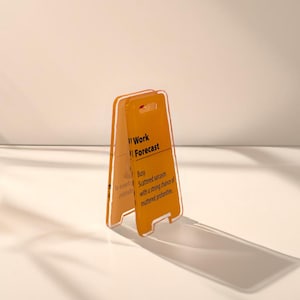 May include: A yellow plastic sign with the text "Work Forecast" and the description "Busy. Scattered sarcasm with a strong chance of muttered profanities."