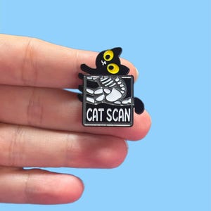 May include: A black enamel pin featuring a cartoon cat with yellow eyes peeking over a rectangular frame. The frame displays a skeleton and the words "CAT SCAN" in white text on a black background. The pin is held against a light blue backdrop.