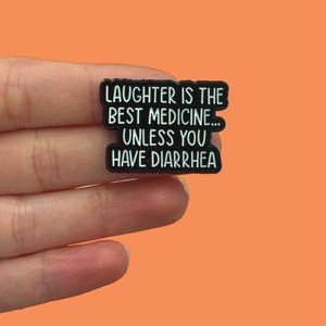 May include: A black enamel pin with the text "LAUGHTER IS THE BEST MEDICINE... UNLESS YOU HAVE DIARRHOEA" in white lettering. The pin is held against an orange background.