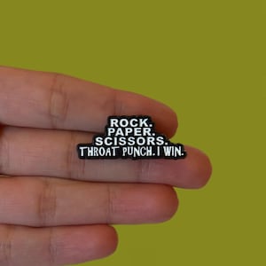 May include: A black rectangular pin with white text that reads "ROCK. PAPER. SCISSORS. THROAT PUNCH. I WIN." The pin is held in a hand against a green background.