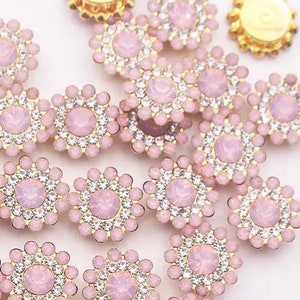 May include: Close-up of several flower-shaped embellishments. Each has a light pink center stone, surrounded by clear rhinestones and a gold-colored metal base. The embellishments are scattered across a white surface.